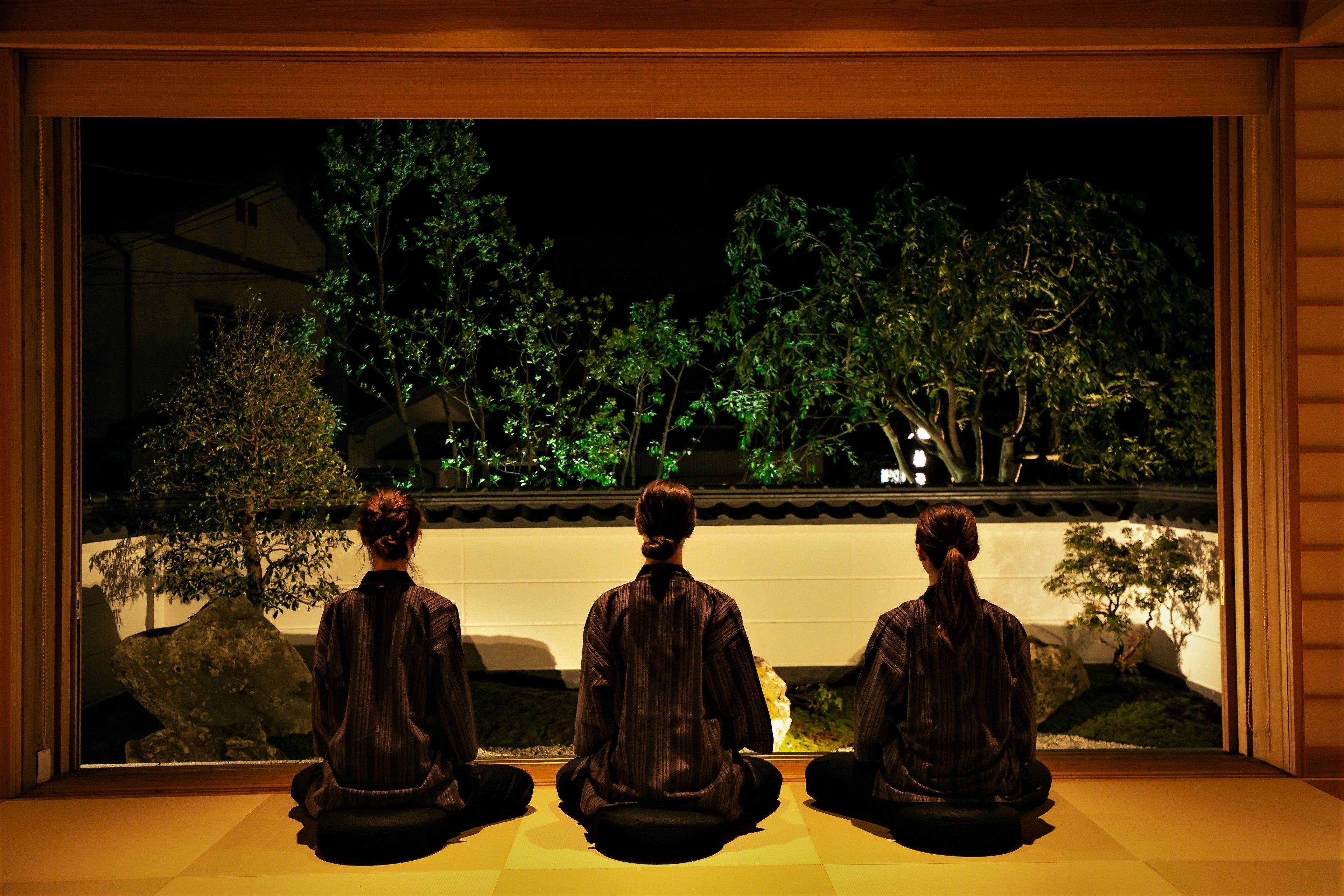 Three individuals seated in a room with a view of a garden at night.