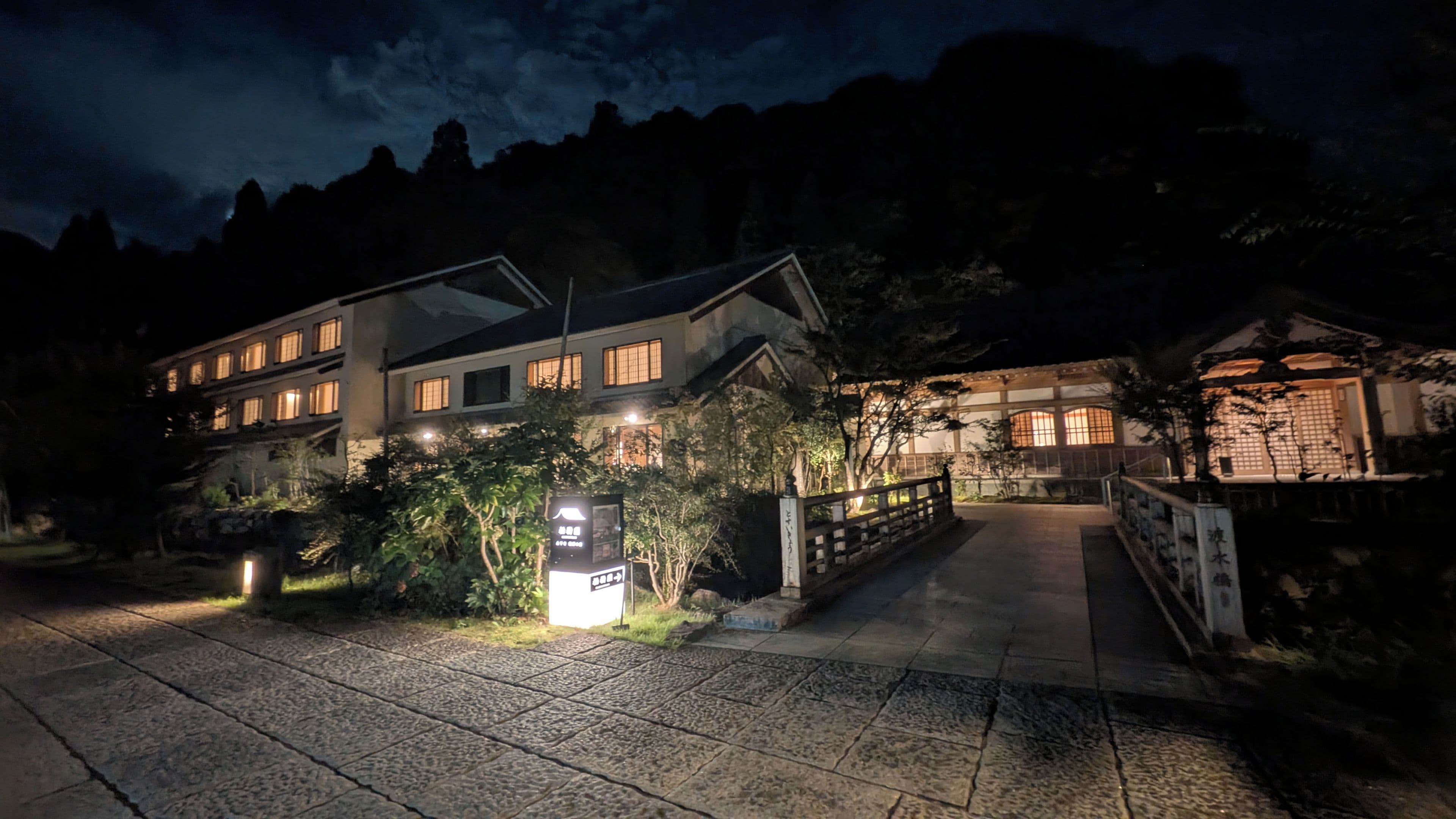 Un hôtel à plusieurs étages, de style architectural japonais traditionnel, doté d’une entrée bien éclairée et d’un chemin de pierre menant au bâtiment principal.