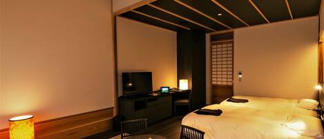 A hotel room with a bed, a desk with a computer, a TV, and a table with a lamp and a chair.