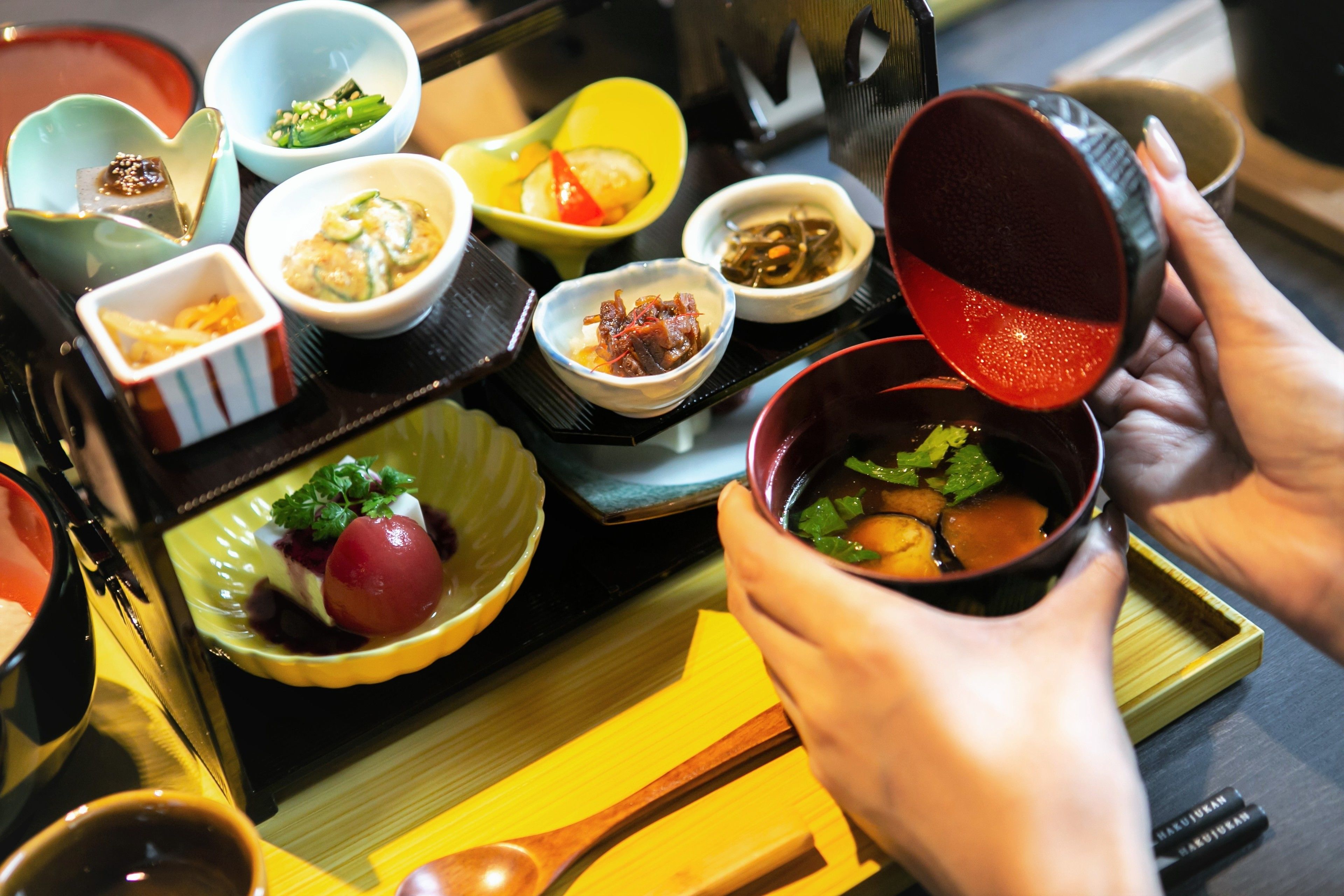 A variety of small dishes with different foods, a person pouring a liquid from a cup, and a tray with a spoon.