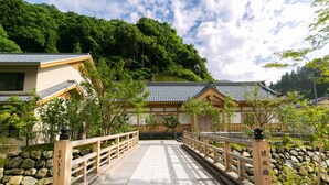 Un pont en bois mène à un bâtiment japonais traditionnel, doté d’un toit en tuiles, et est entouré d’une végétation luxuriante et d’un mur de pierre.