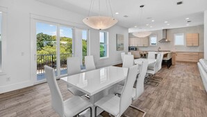Une cuisine moderne avec des meubles blancs, un coin repas doté d’une grande table et de chaises, et de grandes fenêtres offrant une vue sur la verdure.