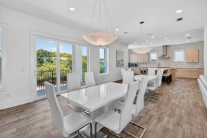A modern kitchen with white cabinets, a dining area with a large table and chairs, and large windows offering a view of greenery.