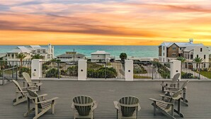 Une terrasse avec des fauteuils Adirondack donnant sur une plage et l’océan, avec des maisons en arrière-plan.