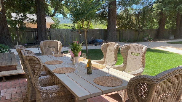 Une terrasse extérieure avec une longue table et des chaises en osier, entourée d’une clôture en bois et d’arbres.