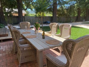 Eine Terrasse im Freien mit einem langen Tisch und Korbstühlen, umgeben von einem Holzzaun und Bäumen.