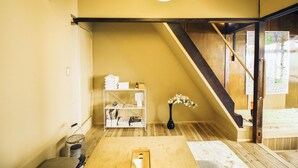 Una habitación con piso de madera, un estante blanco con libros y toallas, un jarrón con flores y una escalera.