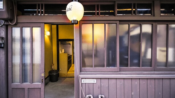 Entrada de una construcción tradicional japonesa, con puerta de madera, un farol y espacio interior visible con mostrador y taburetes.