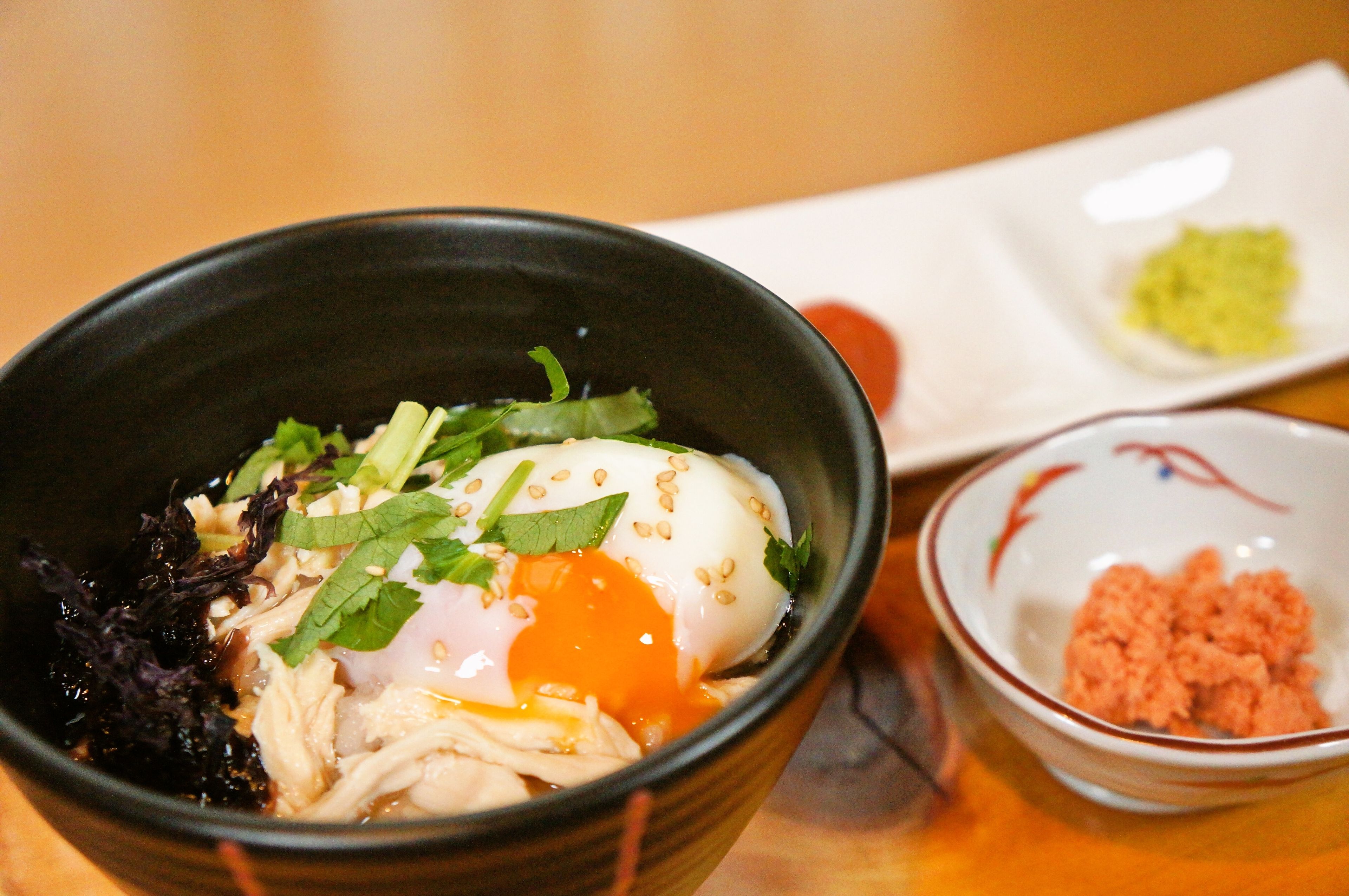 A bowl of food with a poached egg, shredded chicken, and greens, accompanied by a small dish of green sauce and a bowl of pinkish mixture.