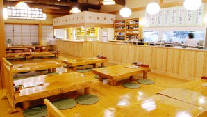 A traditional Japanese restaurant with wooden tables and tatami mats, a counter with a chef, and a menu board with Japanese characters.