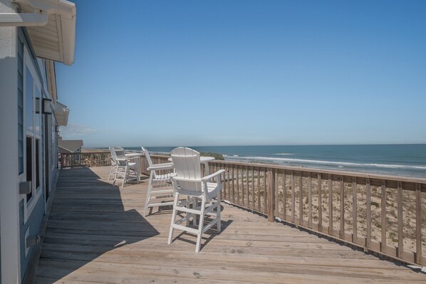 Eine Holzterrasse mit weißen Stühlen und einem Tisch mit Blick auf den Strand.
