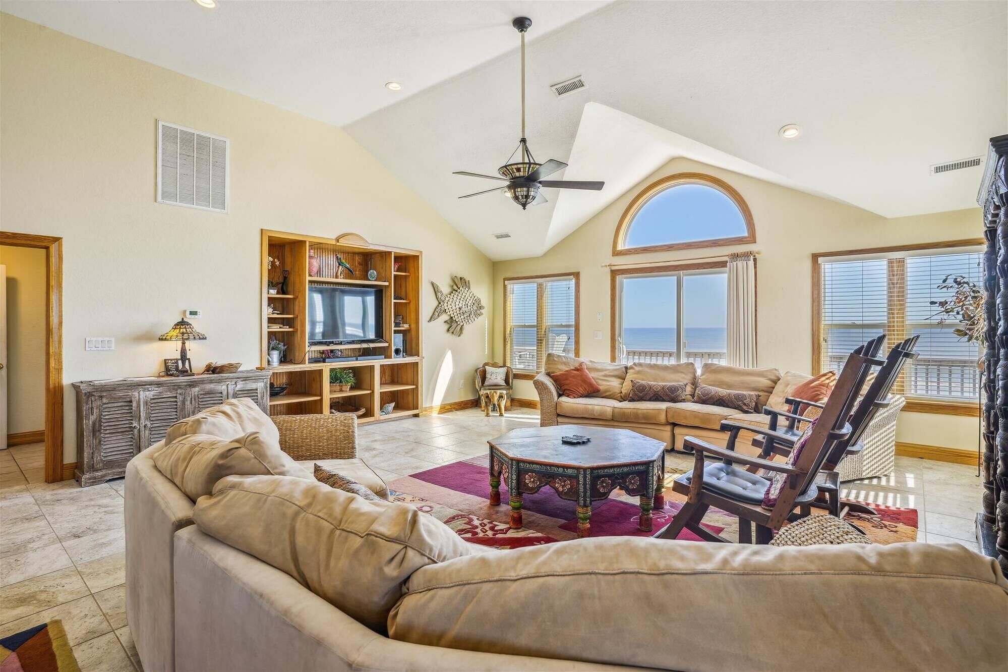 A spacious living room with a high ceiling, a large TV, comfortable seating, and a view of the ocean.