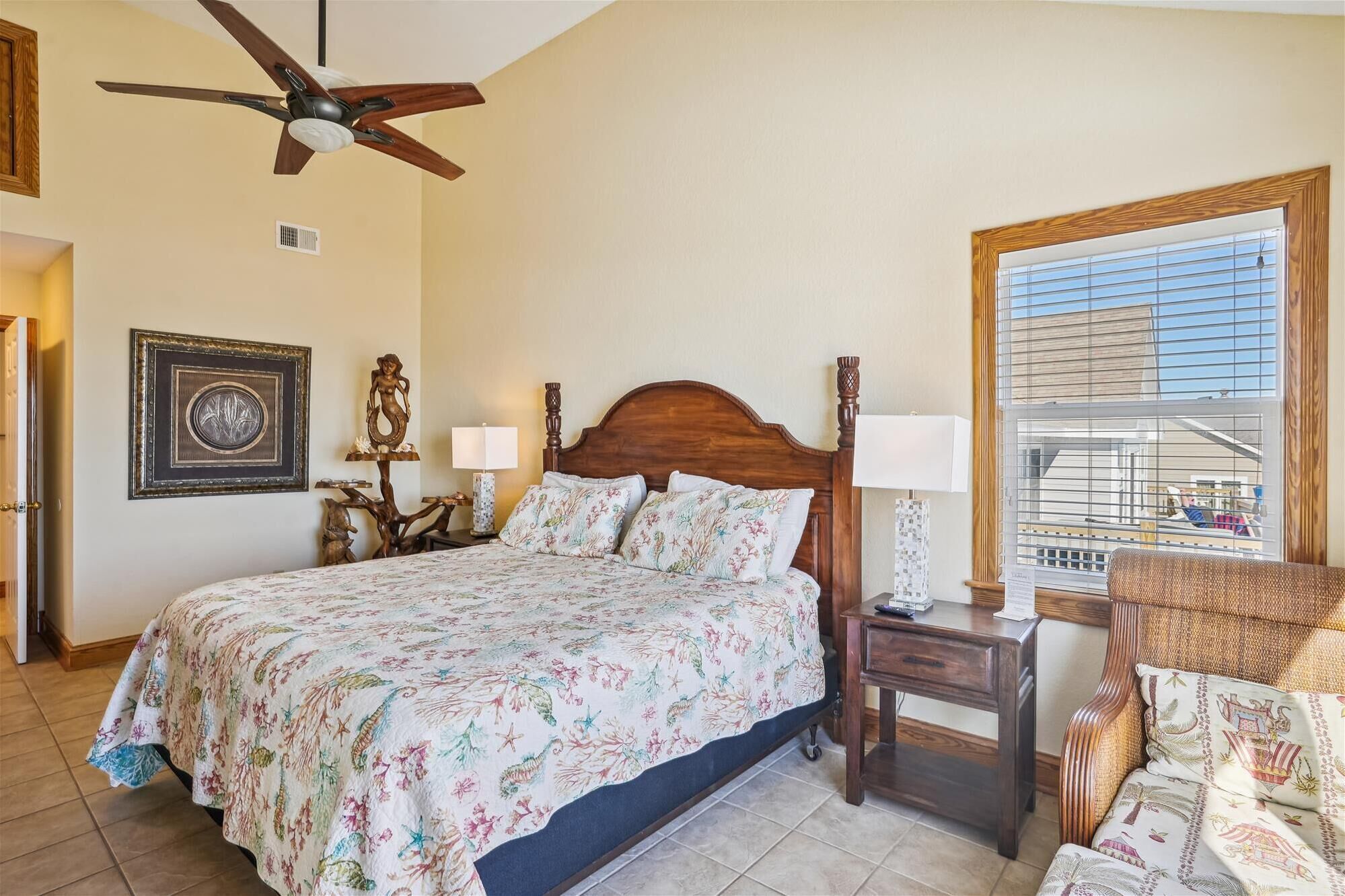 A bedroom with a wooden bed, a nightstand, a lamp, a ceiling fan, a large window with blinds, and a chair.