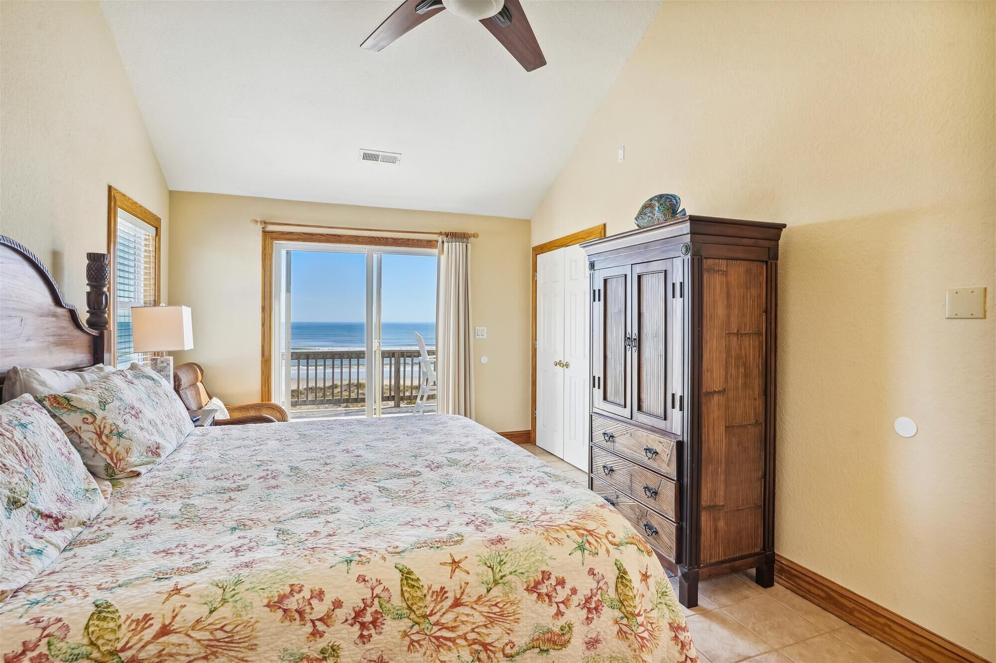 A bedroom with a bed, a nightstand, a dresser, a ceiling fan, and a view of the ocean through a sliding door.
