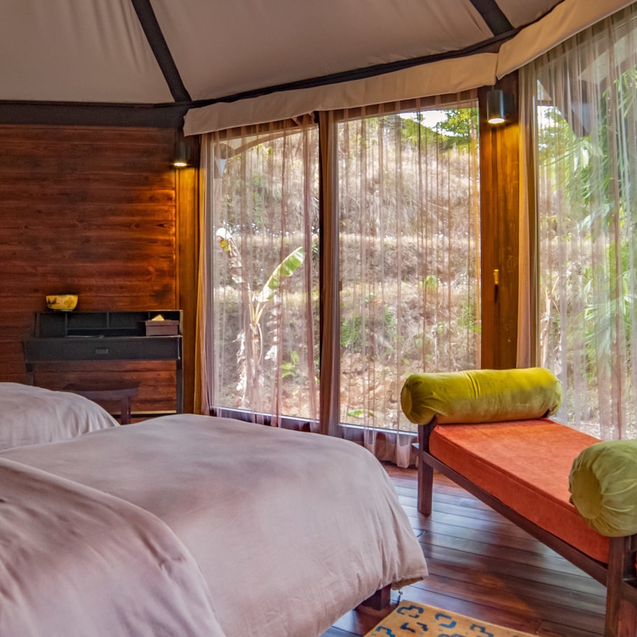 A room with two beds, a wooden bench, and a large window with sheer curtains.
