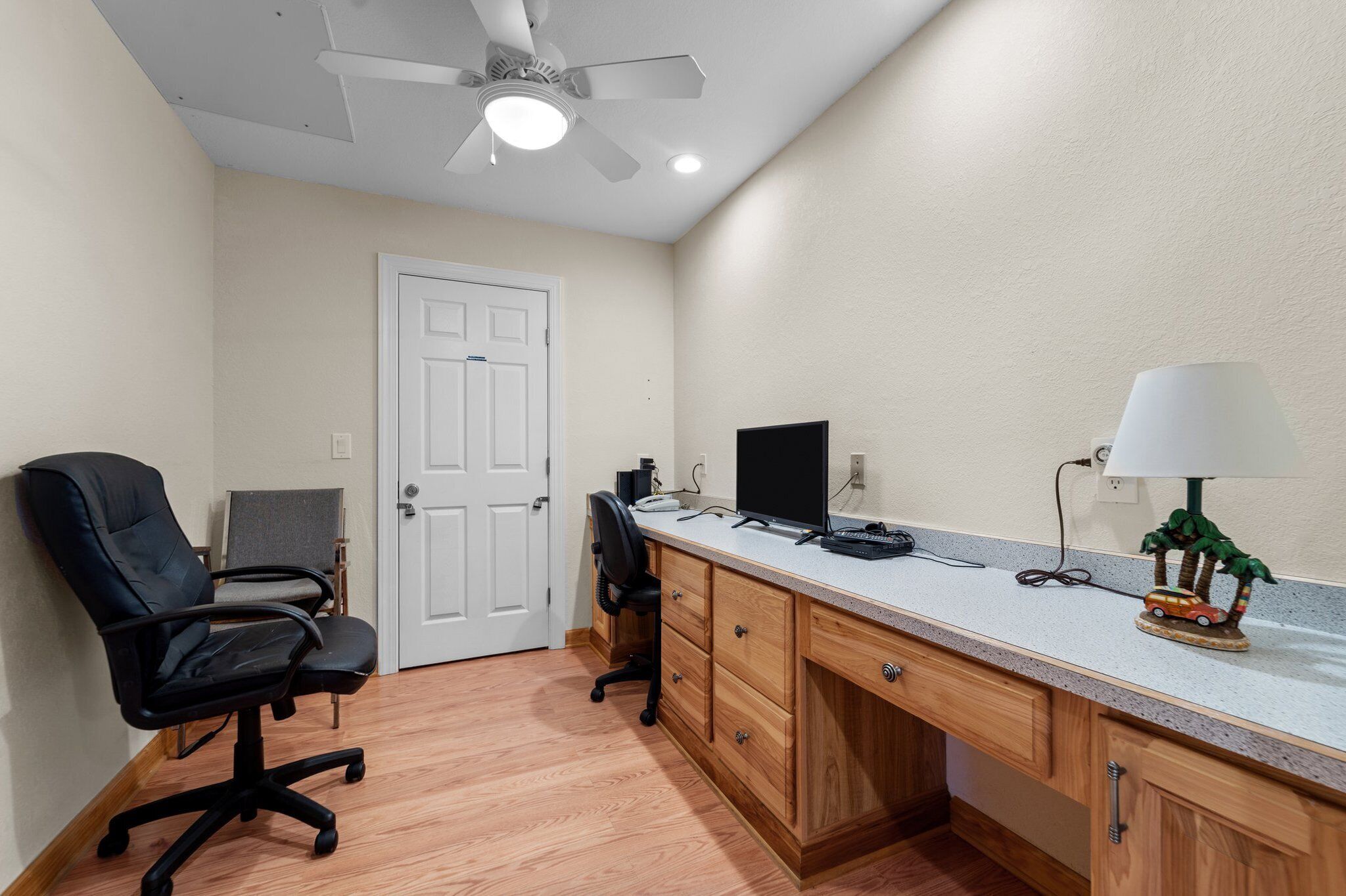 A room with a desk, computer, chair, lamp, and ceiling fan.