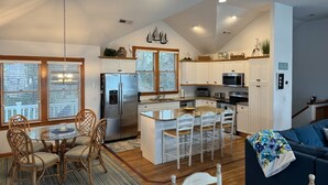 A modern kitchen with a central island, white cabinetry, and a dining area with wicker chairs. - 1289 - Sounds Like Fun, Soundfront Corolla | Pool, Hot Tub & Resort Access (Corolla)