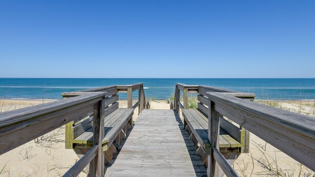 Een houten looppad dat naar een strand leidt, onder een heldere blauwe lucht.