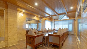 A log cabin-style room with wooden walls and ceiling, a long dining table with chairs, and large windows allowing natural light. - SNOW PEAK RETREAT & COTTAGES - MANALI (Centrally Heated) (Manali)