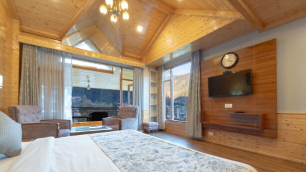 A spacious bedroom with a large bed, a TV mounted on the wall, and a view of the mountains through the windows. - SNOW PEAK RETREAT & COTTAGES - MANALI (Centrally Heated) (Manali)