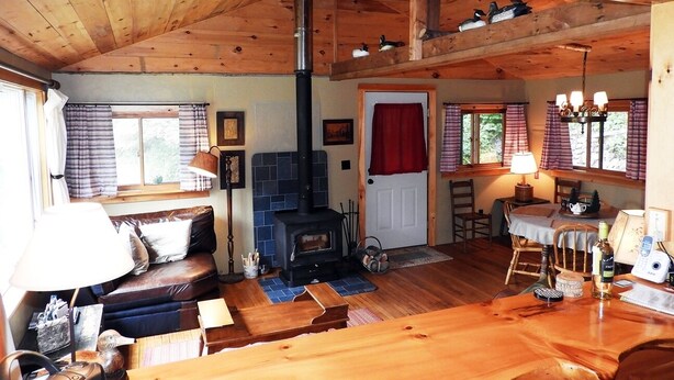 Ein gemütliches Blockhaus-Interieur mit Holzbalkendecke, Kamin, Essbereich und einer Sitzecke mit Sofa und Tisch.