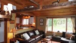 Ein gemütliches Blockhaus-Interieur mit Holzbalkendecke, einem braunen Ledersofa, einem Holztisch und einer Küchenzeile mit Blick auf Grünflächen.
