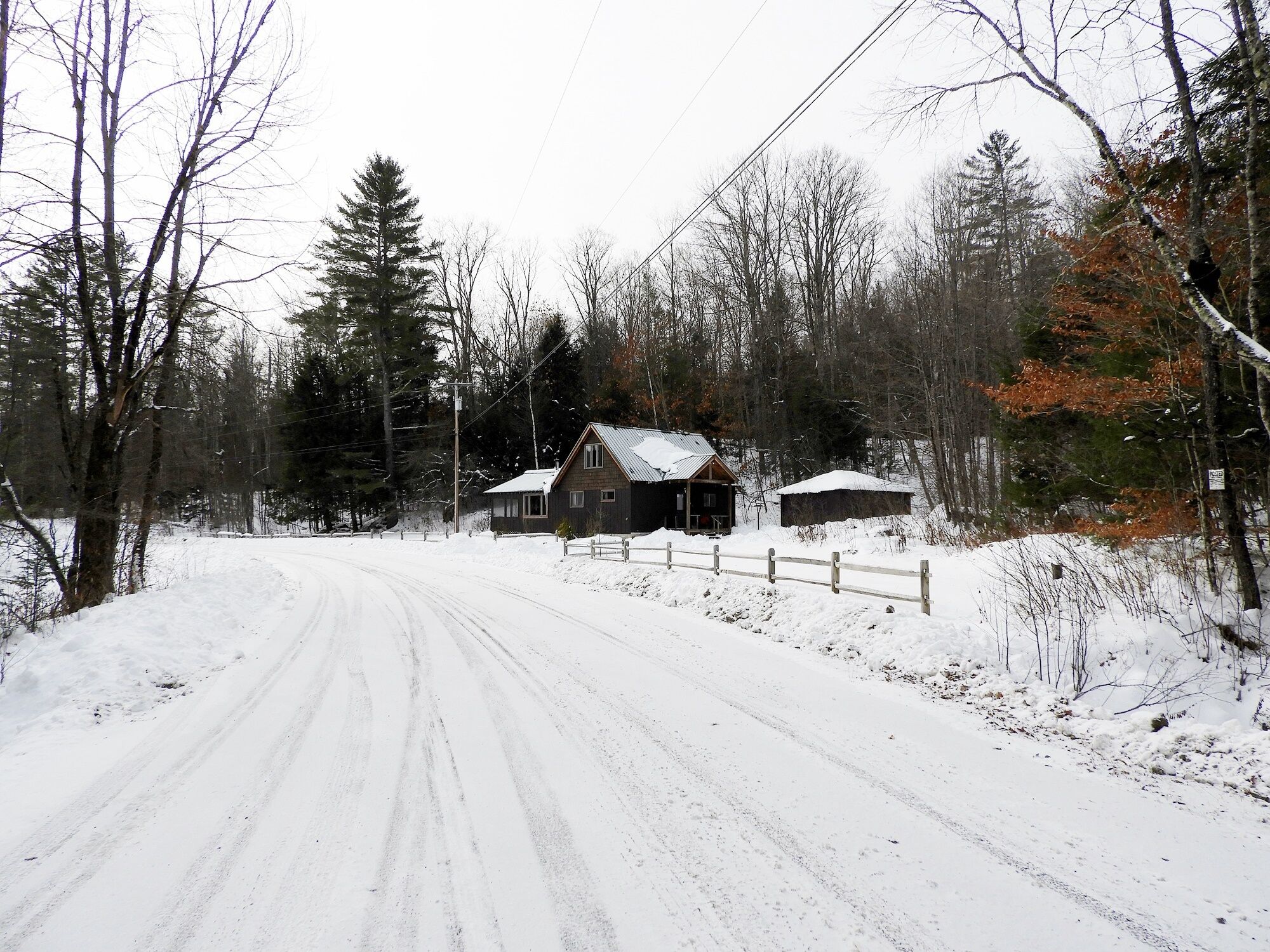 A snow-covered rural road with a wooden cabin and a smaller shed in the background.