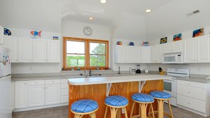 Une cuisine moderne avec des meubles blancs, un îlot central en bois doté de tabourets, une grande fenêtre et un four à micro-ondes.