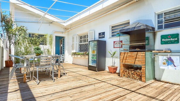 Un patio exterior con cubierta de madera, una mesa con sillas, un refrigerador, una parrilla y plantas en macetas.