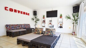 Una sala de estar con un sofá y una cama hecha de palés de madera, un televisor de pantalla plana, y estantes con plantas y adornos.