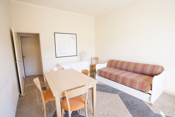 Una habitación pequeña y bien iluminada con una mesa y sillas de madera, un sofá blanco y un dibujo arquitectónico enmarcado en la pared.