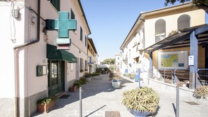 Eine schmale Straße mit Gebäuden an beiden Seiten, darunter eine Apotheke mit einem grünen Vordach. Im Vordergrund befinden sich Kübelpflanzen und ein Metall-Bollard.
