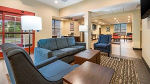 A hotel lobby with a sofa, armchair, and a small table. There is a reception desk, a dining area, and a TV mounted on the wall.
