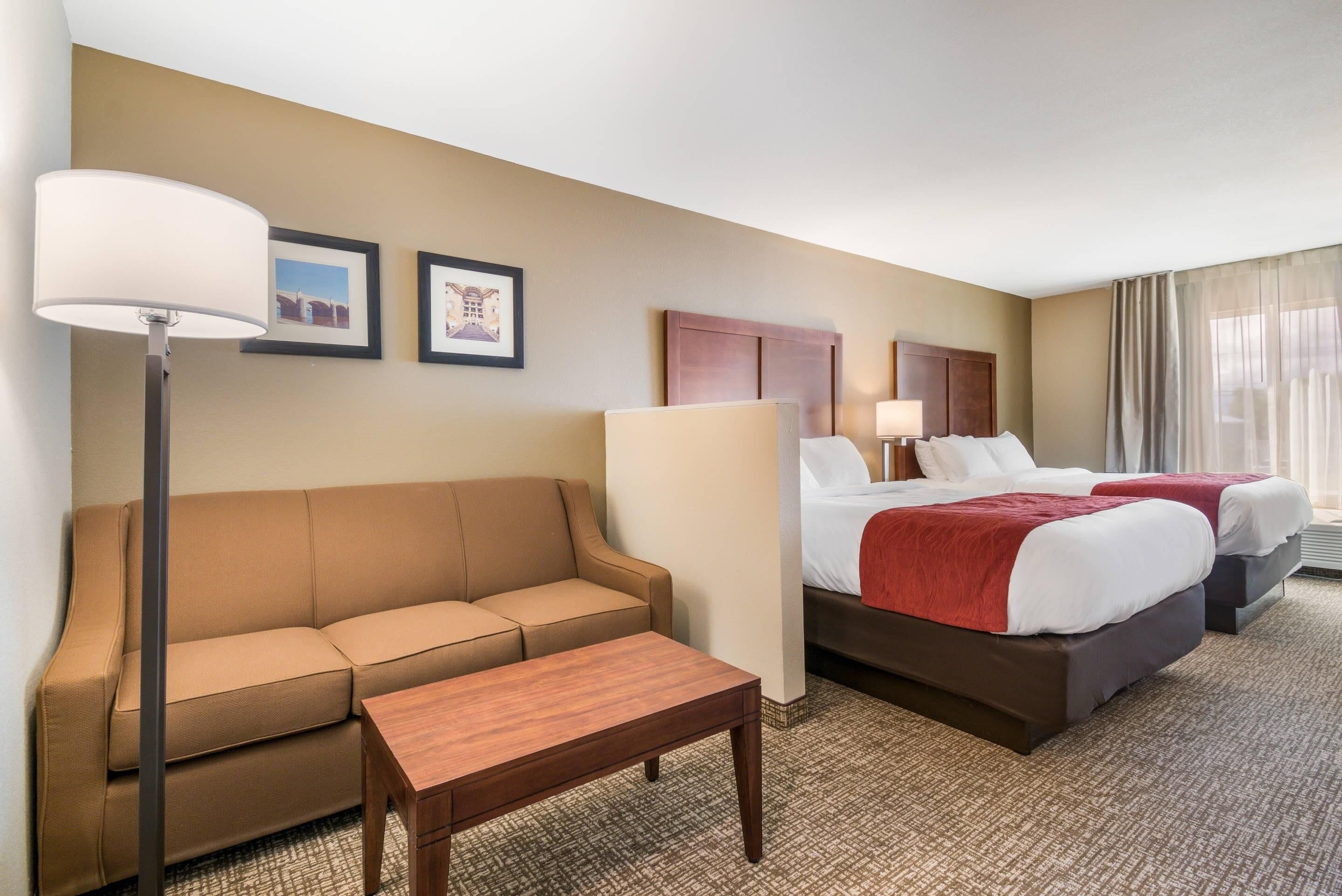 A hotel room with a sofa, two beds, a nightstand, a lamp, and framed pictures on the wall.