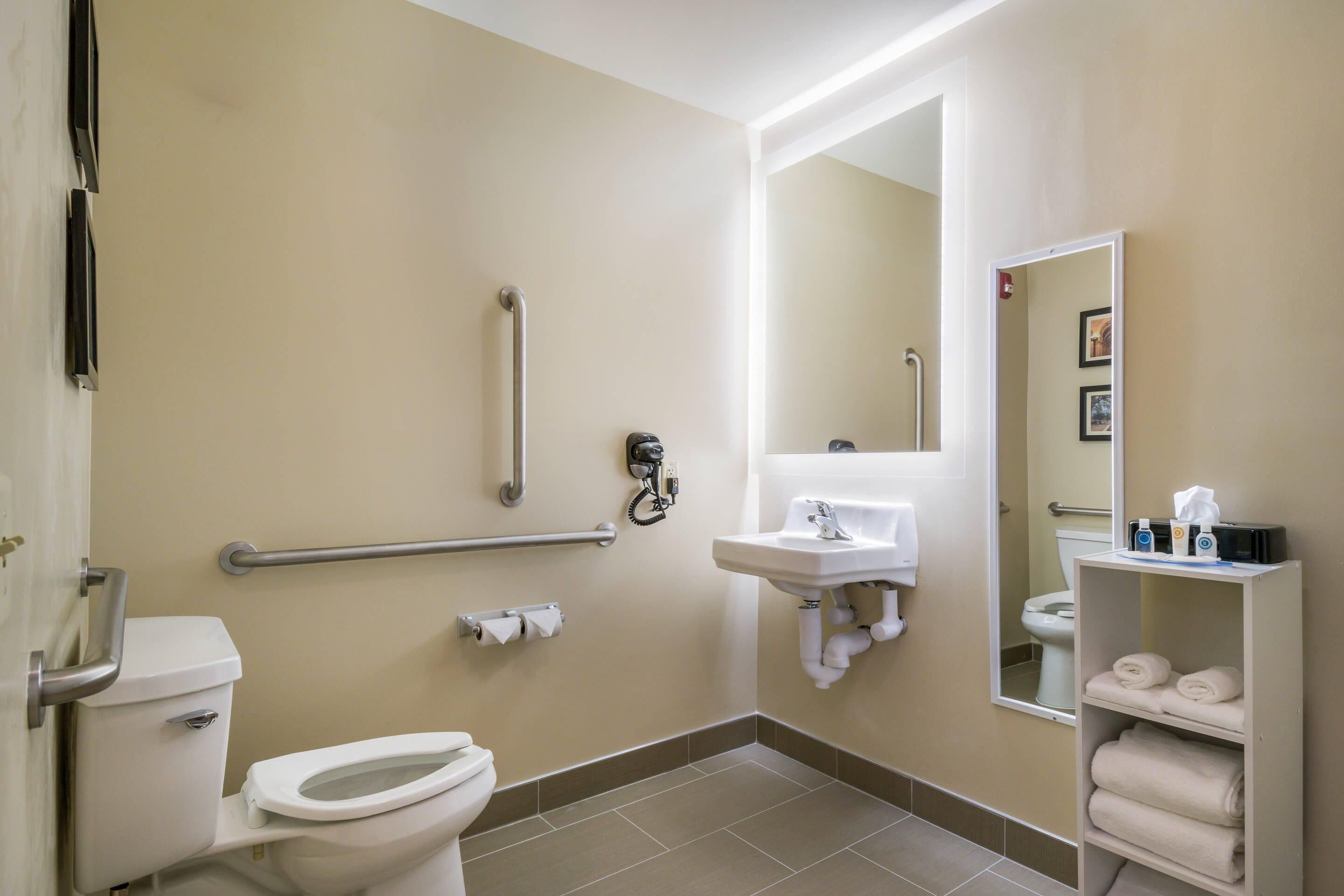 A bathroom with a toilet, handrails, a sink, a mirror, and a shelf with towels.