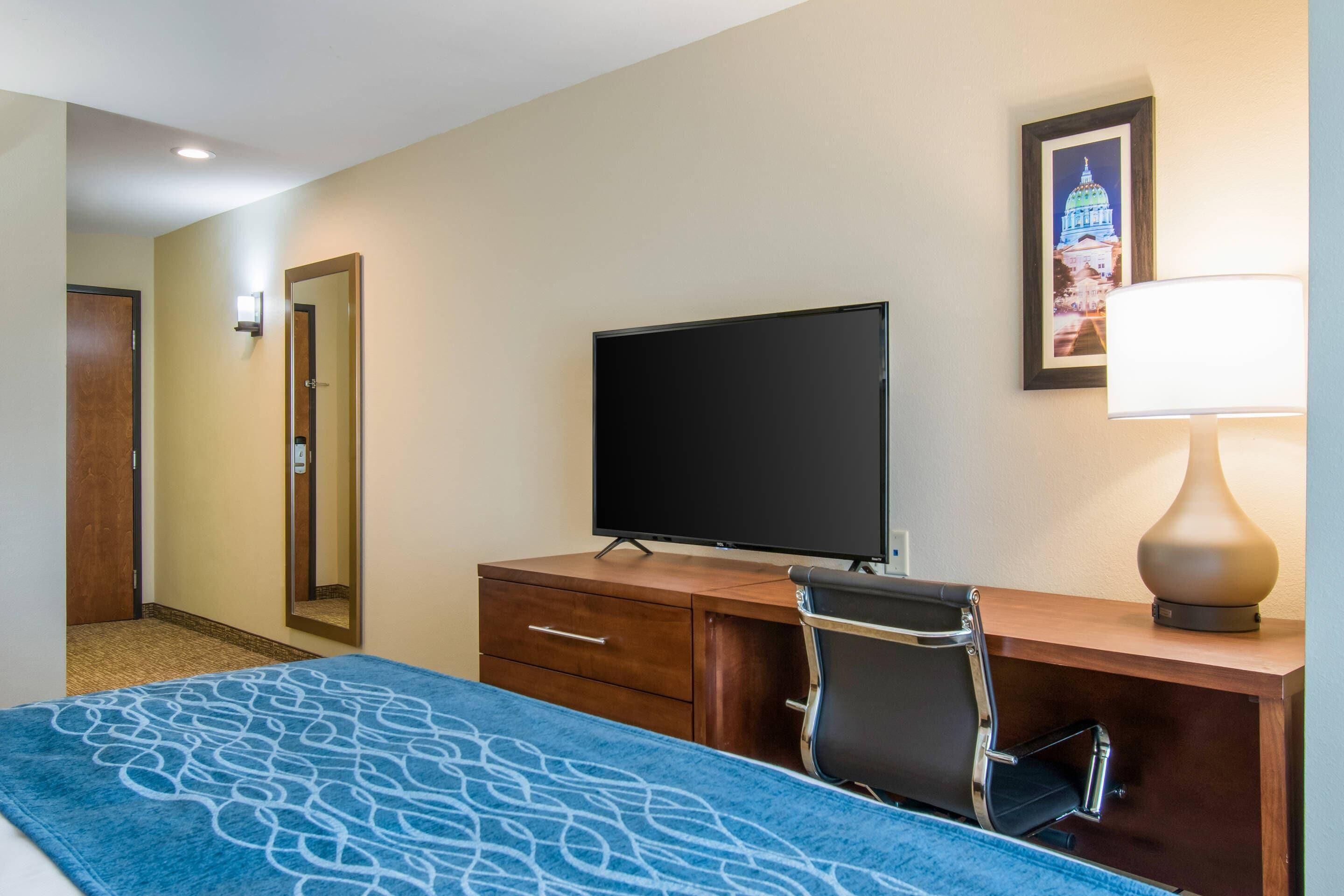 A hotel room with a bed, a desk with a television, a chair, a lamp, and a framed picture on the wall.