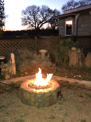 Una chimenea de piedra con llamas, rodeada por un jardín con diversas plantas y una valla de madera.