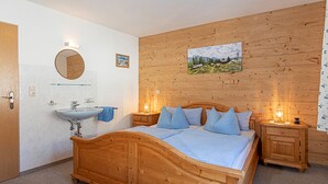 Un dormitorio con cama de madera, una mesita de noche de madera, una mesilla auxiliar, un espejo redondo, cabecero de madera, un panel de pared de madera, un lavabo blanco, una toalla y una alfombra mullida.