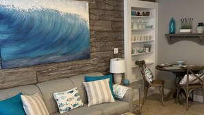 Un salon avec un canapé gris, des coussins turquoise, une table basse en bois, un grand tableau représentant une vague et un ventilateur de plafond.