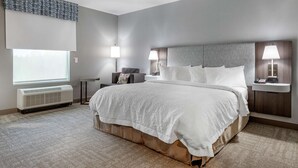 A modern hotel room with a large bed, two bedside tables with lamps, a chair, a window with blinds, and an air conditioning unit. - Hampton Inn Camden (Camden)