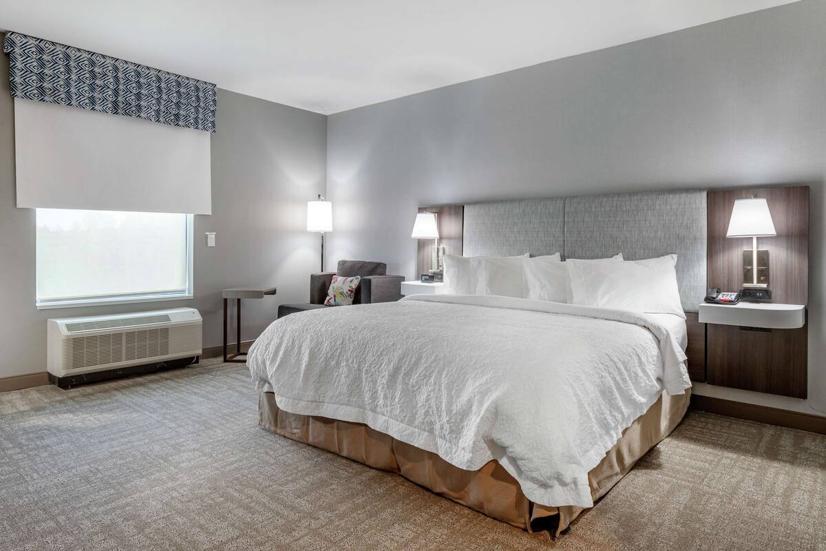 A modern hotel room with a large bed, two bedside tables with lamps, a chair, a window with blinds, and an air conditioning unit.