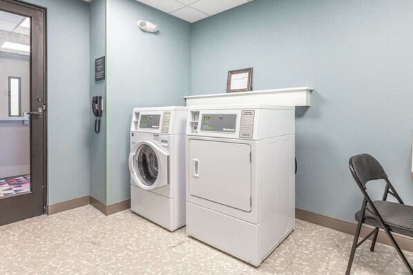 A laundry room with two front-loading washers and dryers, a phone, a shelf with a framed certificate, and a folding chair.