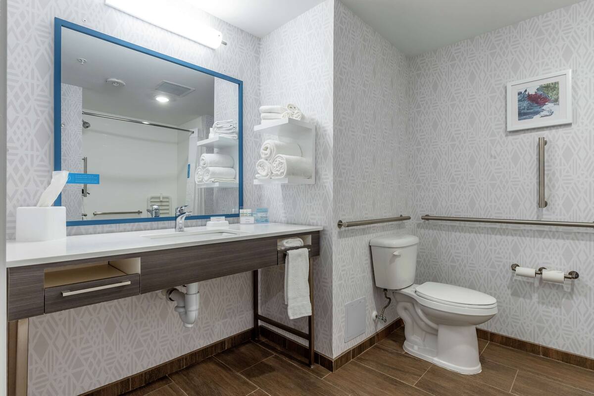 A modern bathroom with a large mirror, a sink with a towel rack, a toilet, and a handrail for accessibility.
