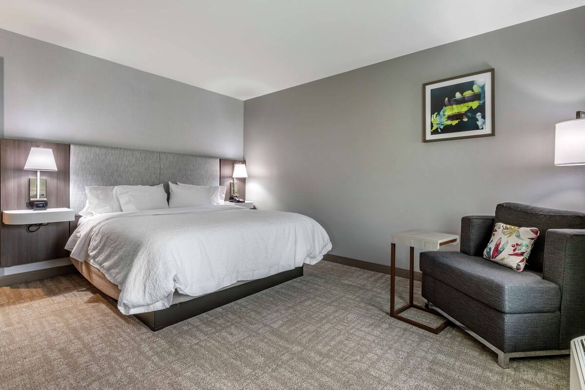 A hotel room with a bed, two bedside lamps, a gray armchair, a small table, and a framed picture on the wall.