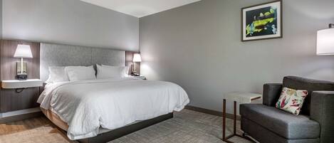A hotel room with a bed, two bedside lamps, a gray armchair, a small table, and a framed picture on the wall.