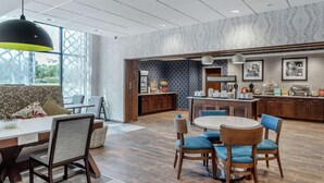 A modern hotel lobby with a dining area, a kitchen, and a seating area with a sofa and chairs.