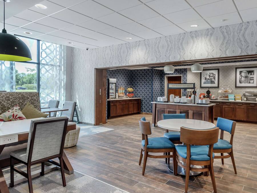 A modern hotel lobby with a dining area, a kitchen, and a seating area with a sofa and chairs.