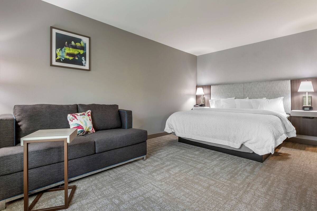 A hotel room with a grey sofa, a small table, a bed with white bedding, and a nightstand with lamps.