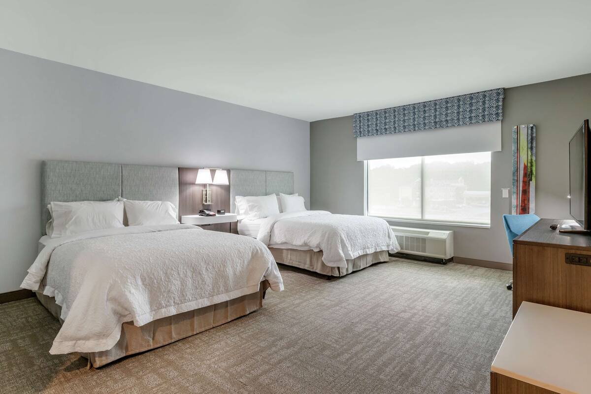A hotel room with two beds, a desk, a chair, a lamp, a window with a view, and a radiator.