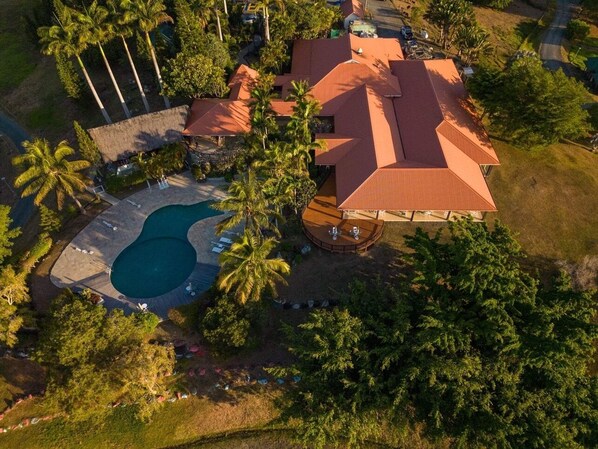 An aerial view of a house with a swimming pool, surrounded by palm trees and a well-maintained lawn.